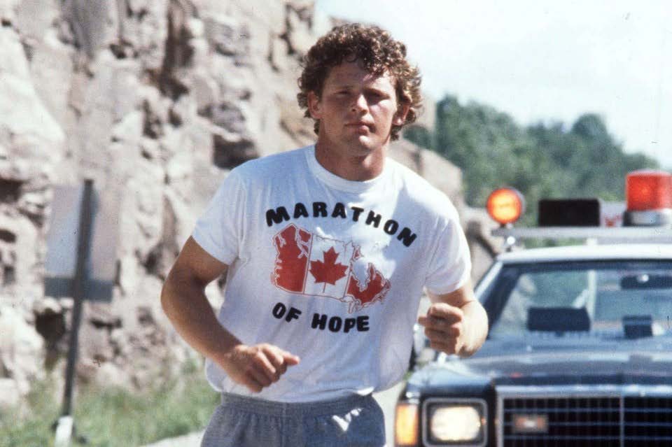 Archival photo of Terry Fox, wearing a t-shirt that reads "Marathon of Hope" running in front of a police escort. 